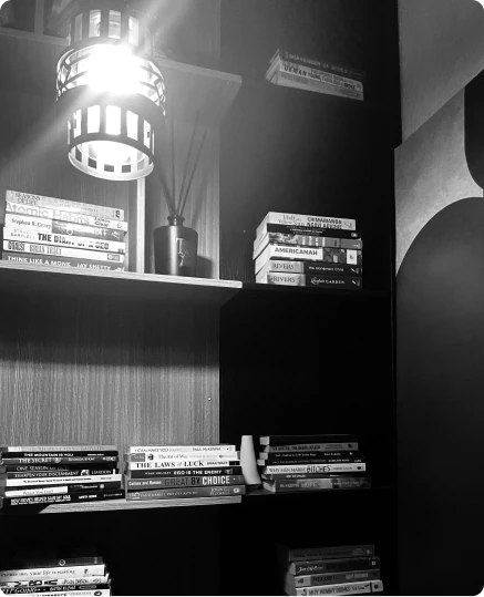 Dark shelves lit by a lamp and filled with stacked books and DVDs, in black and white