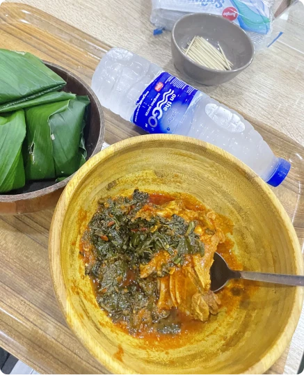 A bowl of leafy stew with a spoon, beside wrapped leaves and a bottle of water
